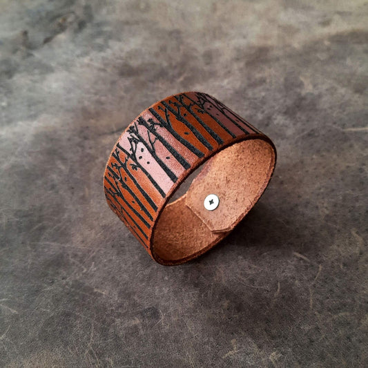 A brown leather cuff bracelet with a tree and branch engravings, displayed on a textured background.