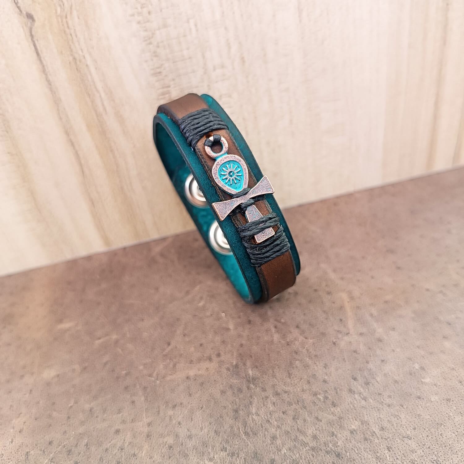 A unisex leather bracelet with an ankh charm, featuring layers of aqua and petrol blue crazy horse leather, accented with a brown leather strip and black waxed cord rivets. It has a stainless steel snap button closure.