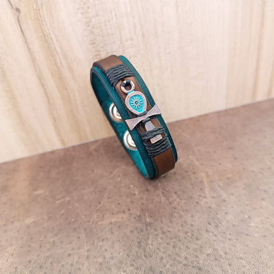 A unisex leather bracelet with an ankh charm, featuring layers of aqua and petrol blue crazy horse leather, accented with a brown leather strip and black waxed cord rivets. It has a stainless steel snap button closure.