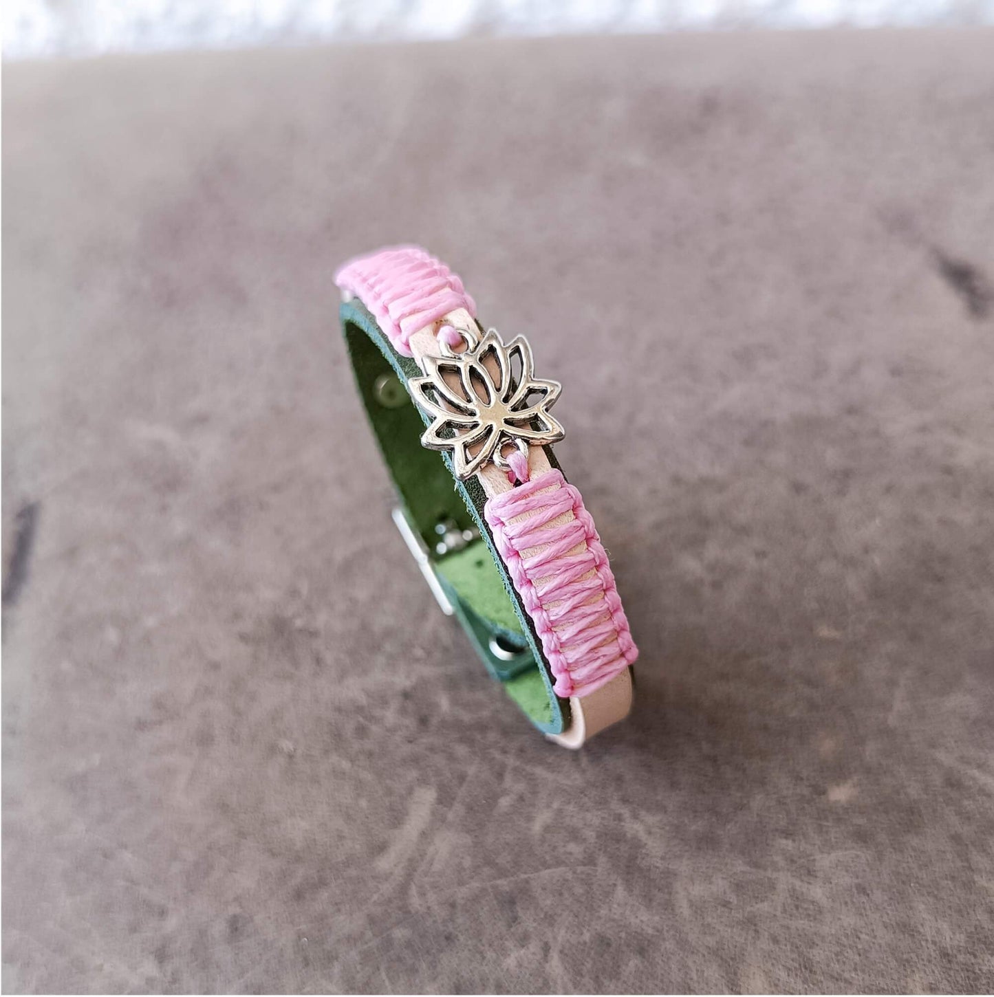 A handmade leather bracelet with pink and green braided straps, featuring a small silver-tone lotus flower charm, resting on a textured brown surface.