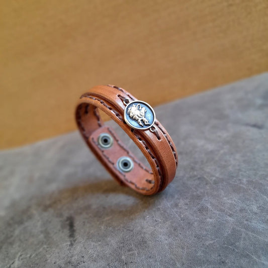 A brown leather bracelet with a layered style, featuring a central boar pendant inspired by Greek mythology, displayed on a textured background.
