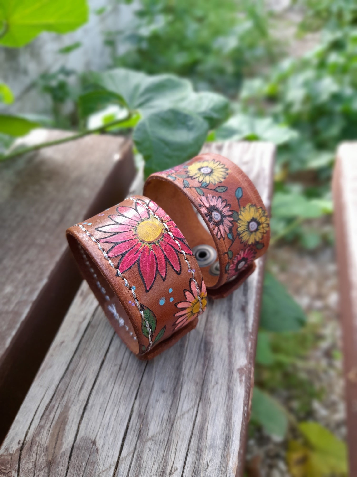 Two handmade and hand painted colorful floral leather cuff bracelets on a wooden bench with green plants on the background.