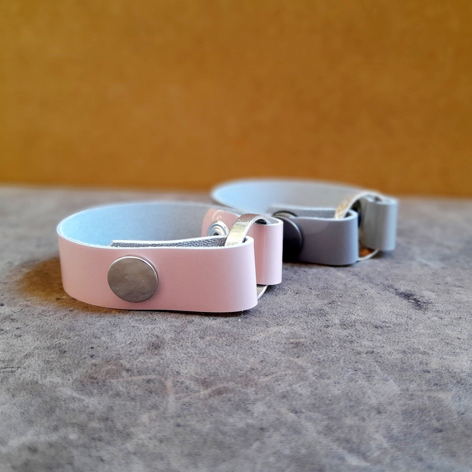 2 handmade glossy leather ringholder bracelets, one soft pink and one soft gray, holding rings.