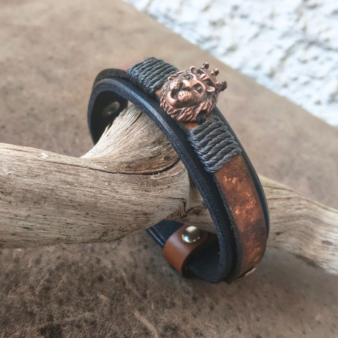 A leather cuff bracelet with a textured dark gray wrap, a copper-toned metal band, and a detailed lion head charm with a crown, resting on a piece of driftwood.