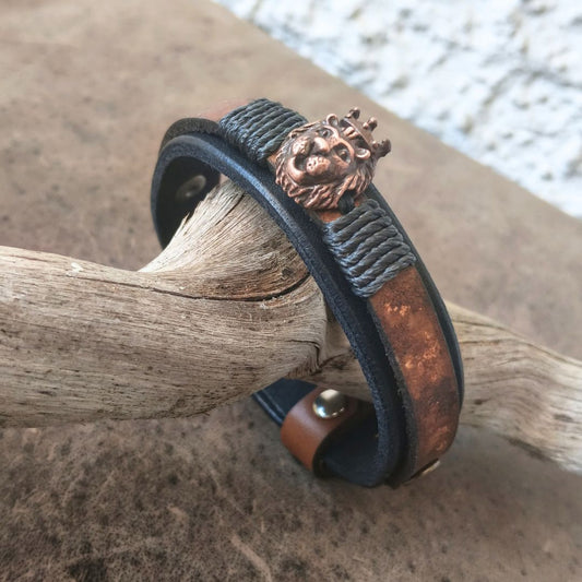 A leather cuff bracelet with a textured dark gray wrap, a copper-toned metal band, and a detailed lion head charm with a crown, resting on a piece of driftwood.