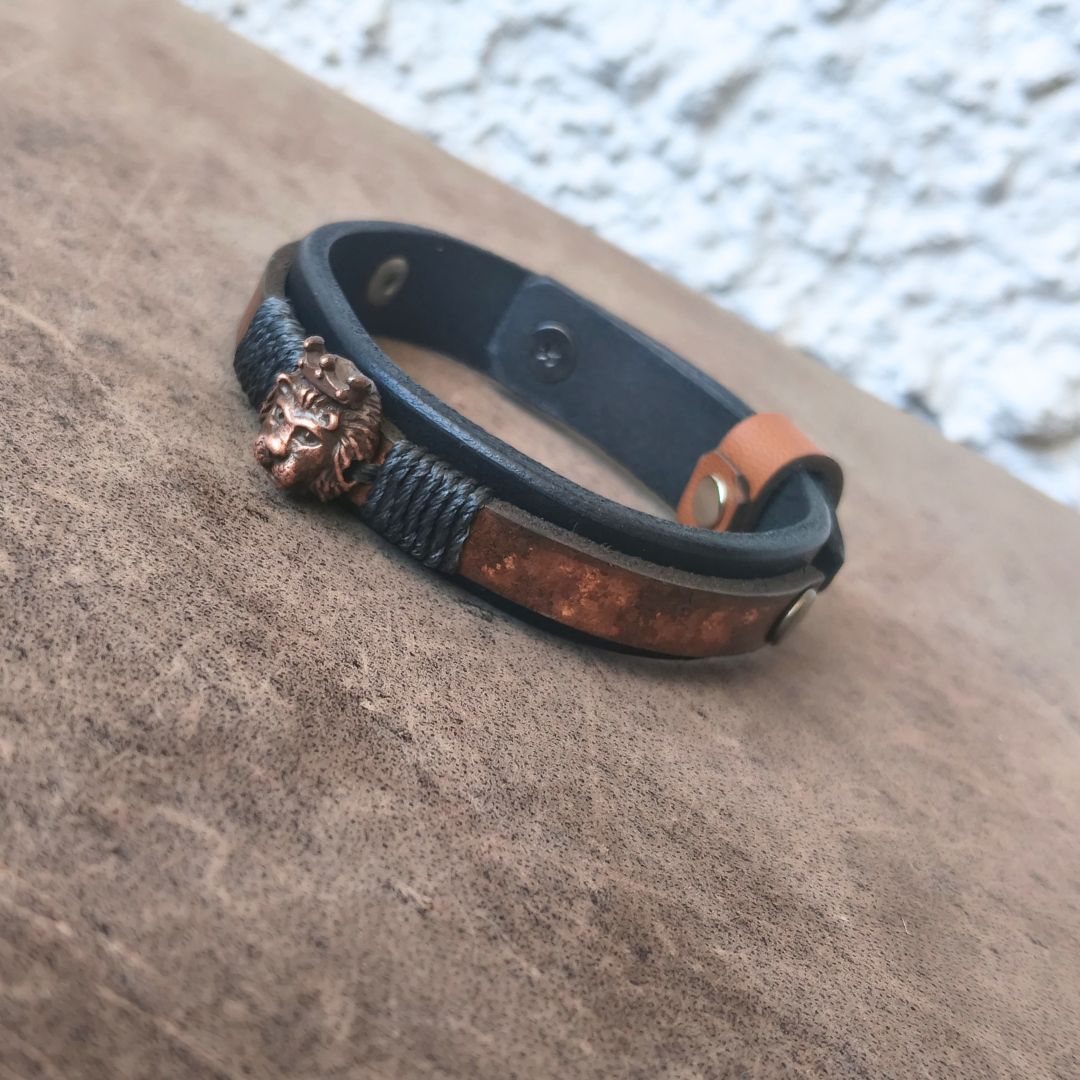 A leather bracelet with a dark brown leather base, a textured black braided band, a copper-toned lion head charm, and a weathered copper inlay, resting on a brown textured surface.