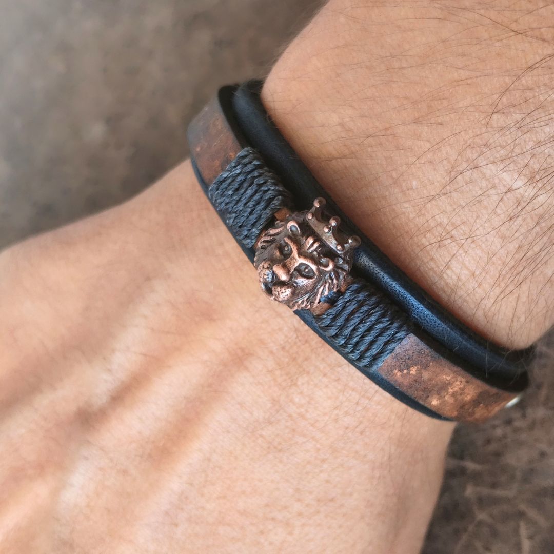 A close-up of a person's wrist wearing a black leather bracelet with a textured braided cord detail and a prominent bronze lion head clasp.