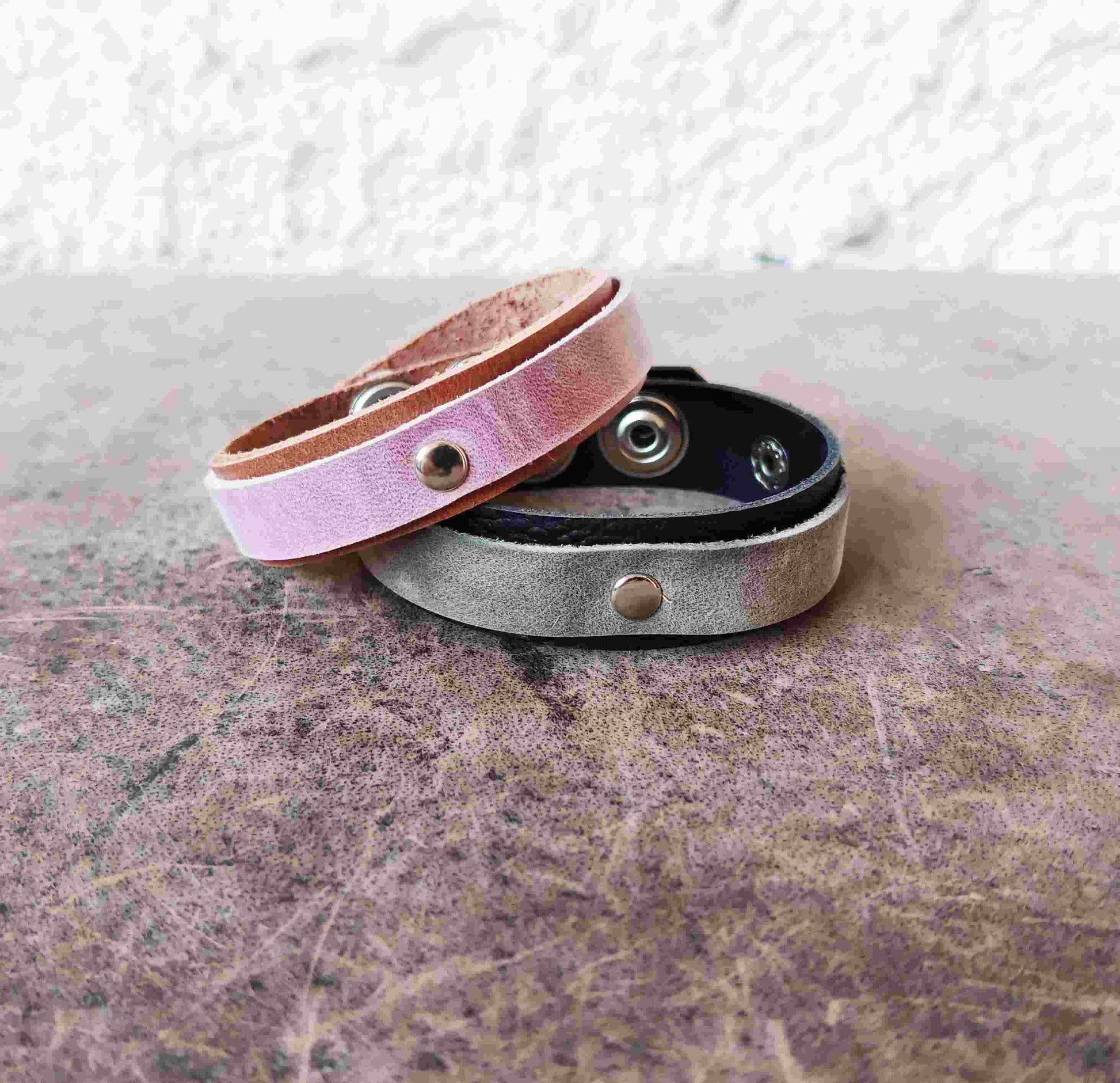 Two slim leather snap bracelets stacked on a textured surface. The top bracelet is light pink with a metallic sheen, and the bottom bracelet is a dark gray with a black interior and a silver-toned snap button.