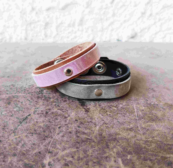 Two slim leather snap bracelets stacked on a textured surface. The top bracelet is light pink with a metallic sheen, and the bottom bracelet is a dark gray with a black interior and a silver-toned snap button.