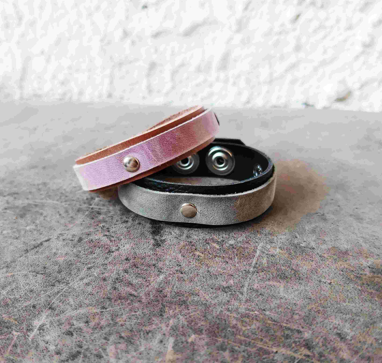 Two leather wristbands stacked on a textured gray surface. The top wristband is a dusty pink leather with a silver snap button, and the bottom wristband is a distressed silver leather with a black interior and silver snap buttons.