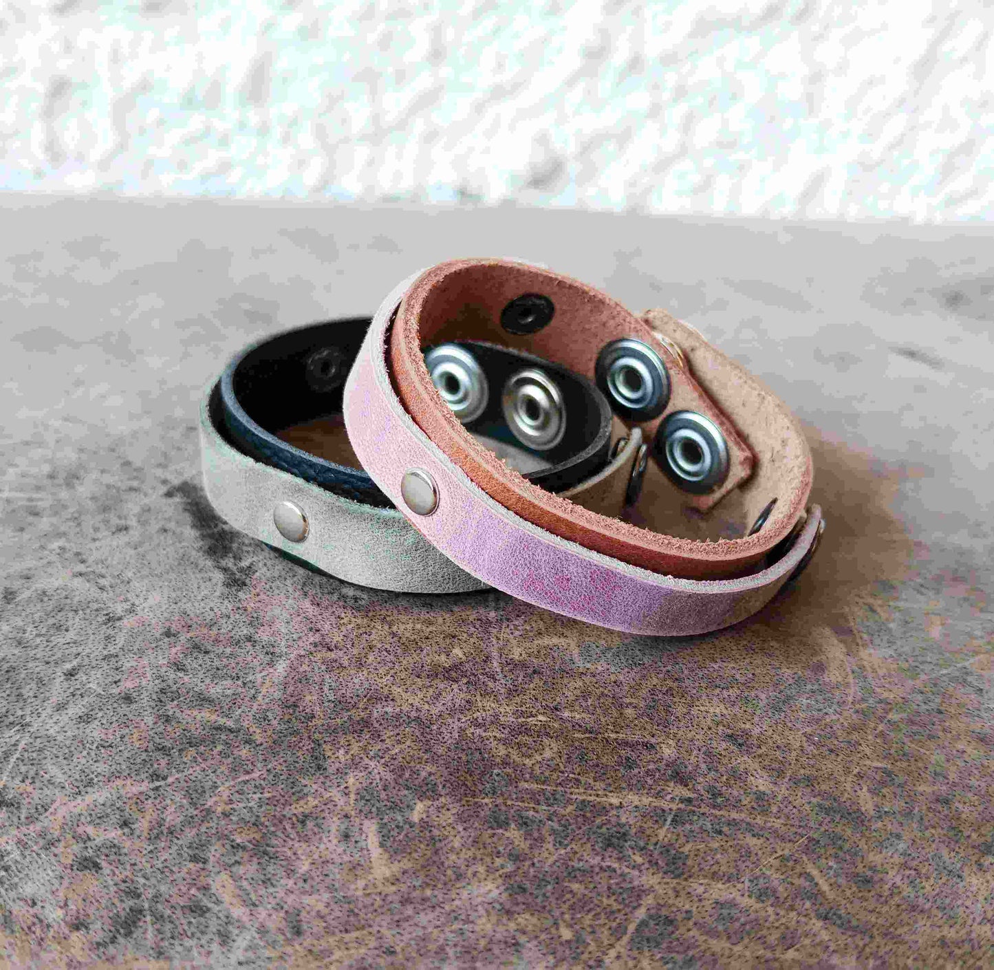 Two leather wristbands with snap-button closures, one in light pink and one in light gray, displayed on a textured gray surface.