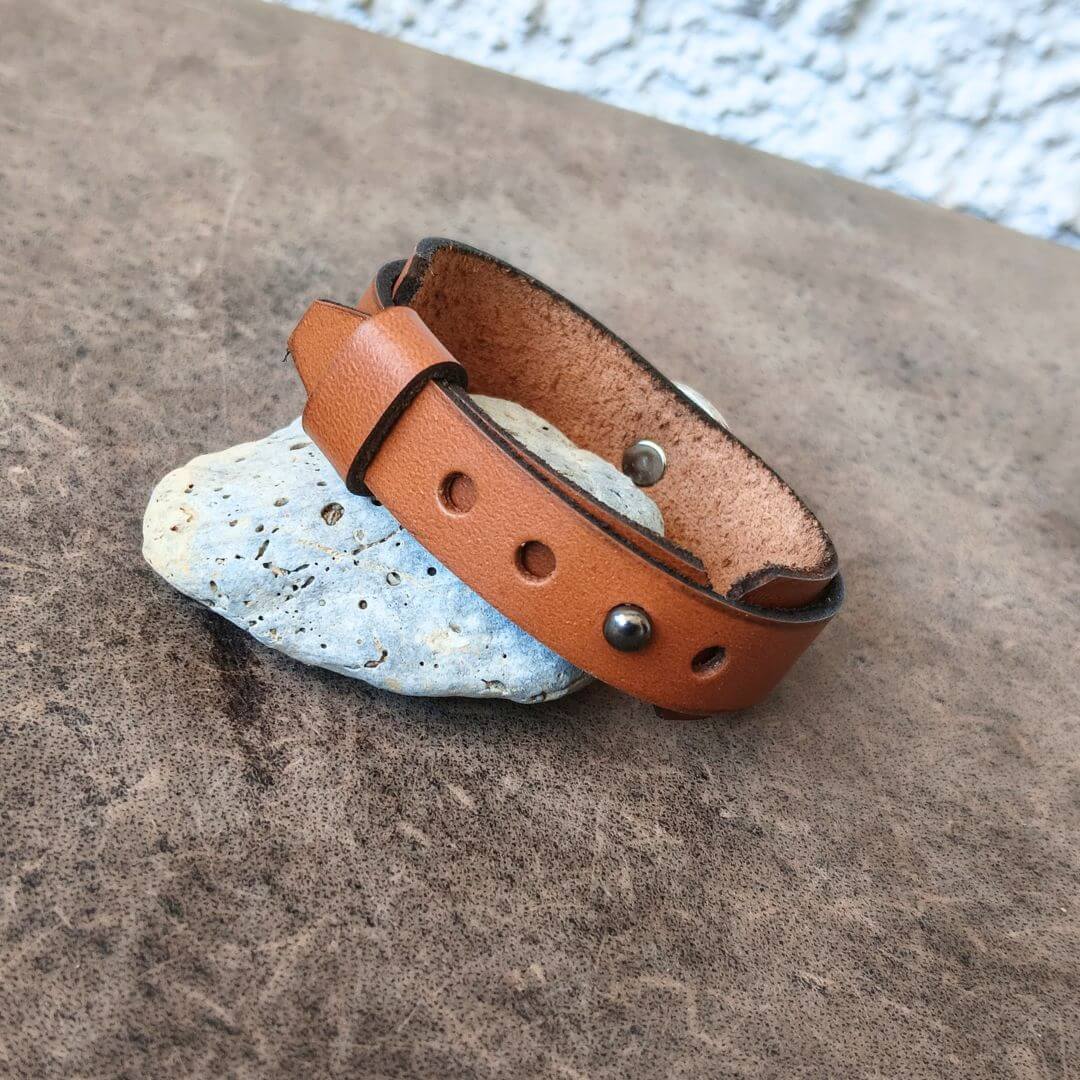 A tan leather adjustable bracelet with silver-tone metal rivets and a buckle, resting on a small light-colored stone against a textured brown surface.