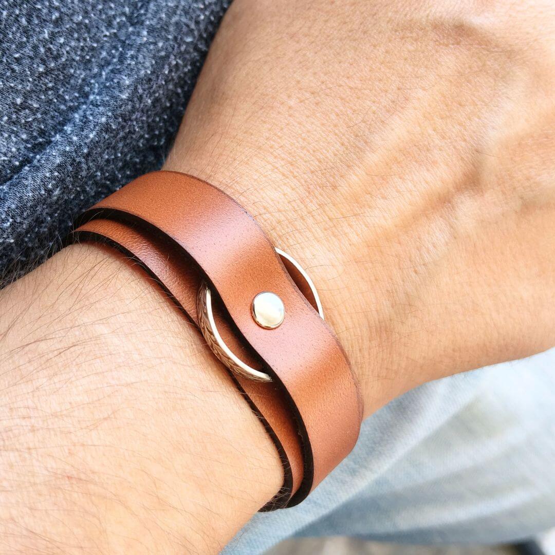A close-up of a person's wrist wearing a brown leather wrap bracelet with a gold-tone metal ring and snap closure.