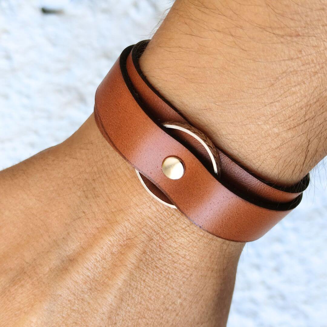 A close-up of a person's wrist wearing a brown leather wrap bracelet with a gold-tone metal ring and snap closure.
