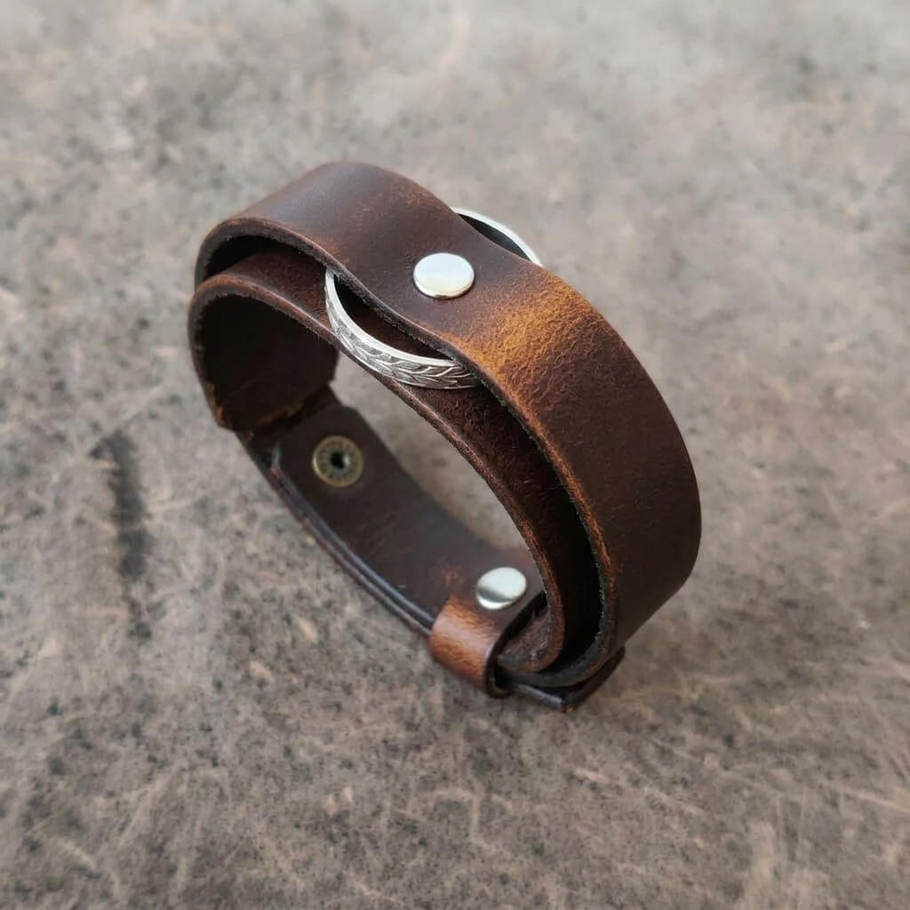 A brown leather wrap bracelet with silver-tone metal rivets and a textured silver band, photographed on a textured gray stone surface.