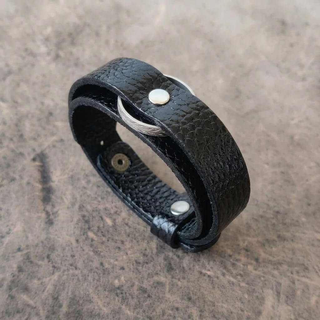 A black textured leather wrap bracelet with silver-toned metal rivets and a thin silver wire accent, displayed on a textured gray surface.