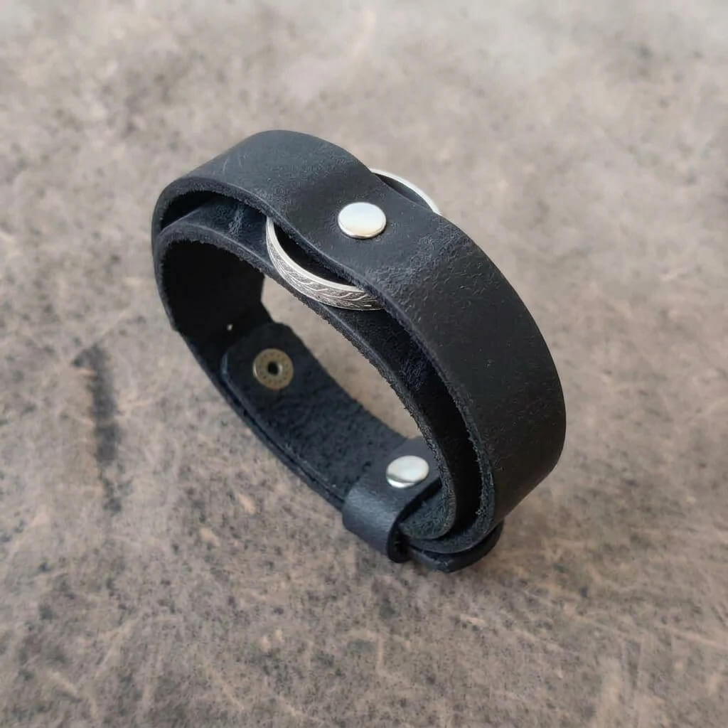 A black leather wrap bracelet with a silver-colored metal cable and silver rivets, displayed on a textured gray surface.