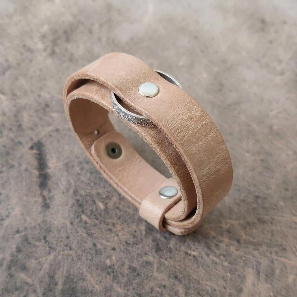 A light tan leather wrap bracelet with silver-tone metal rivets and a silver-tone metal accent, displayed on a textured gray surface.
