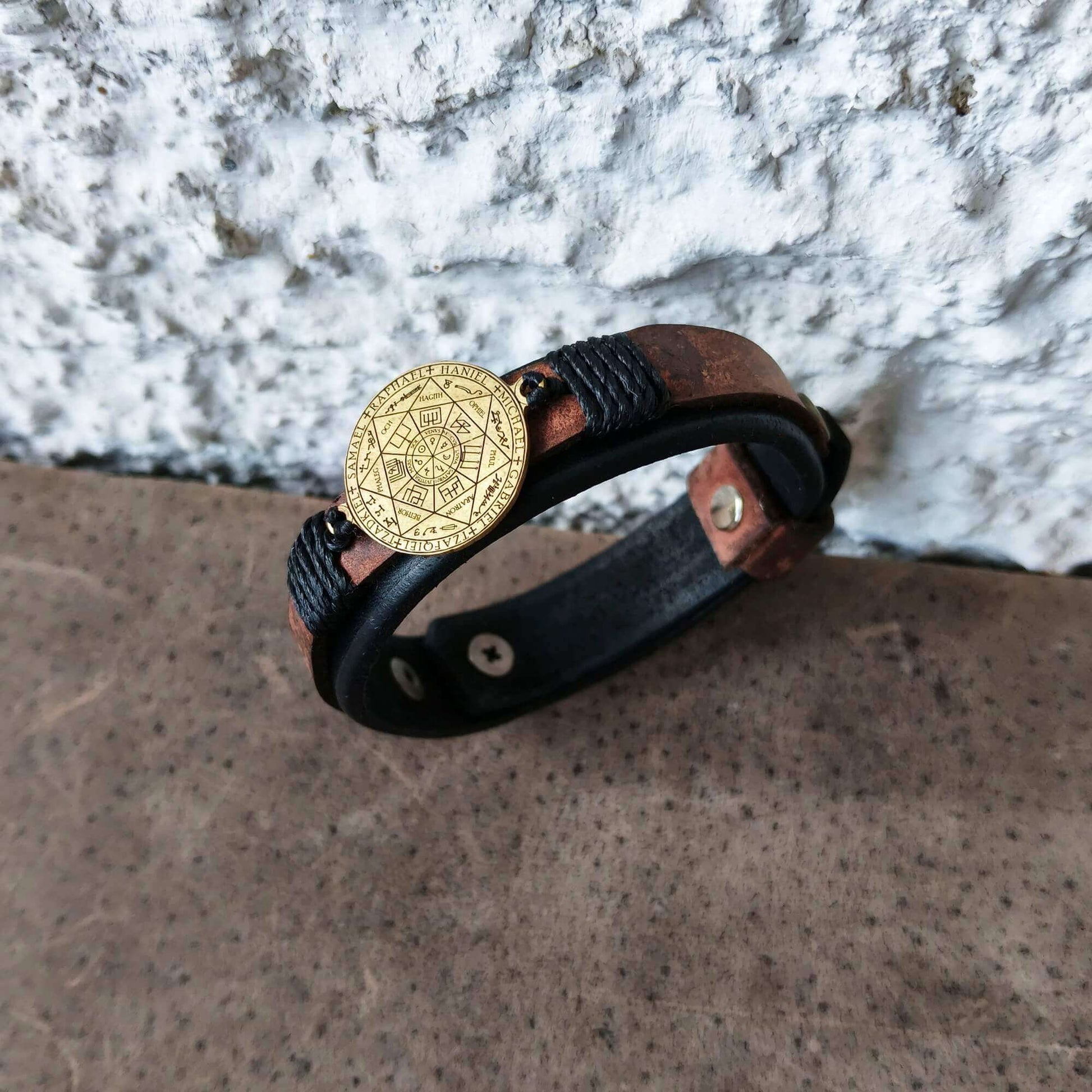 A black leather wrap bracelet with a gold-toned circular 7 Archangels Seal medallion, resting on a textured brown surface against a white stucco wall.