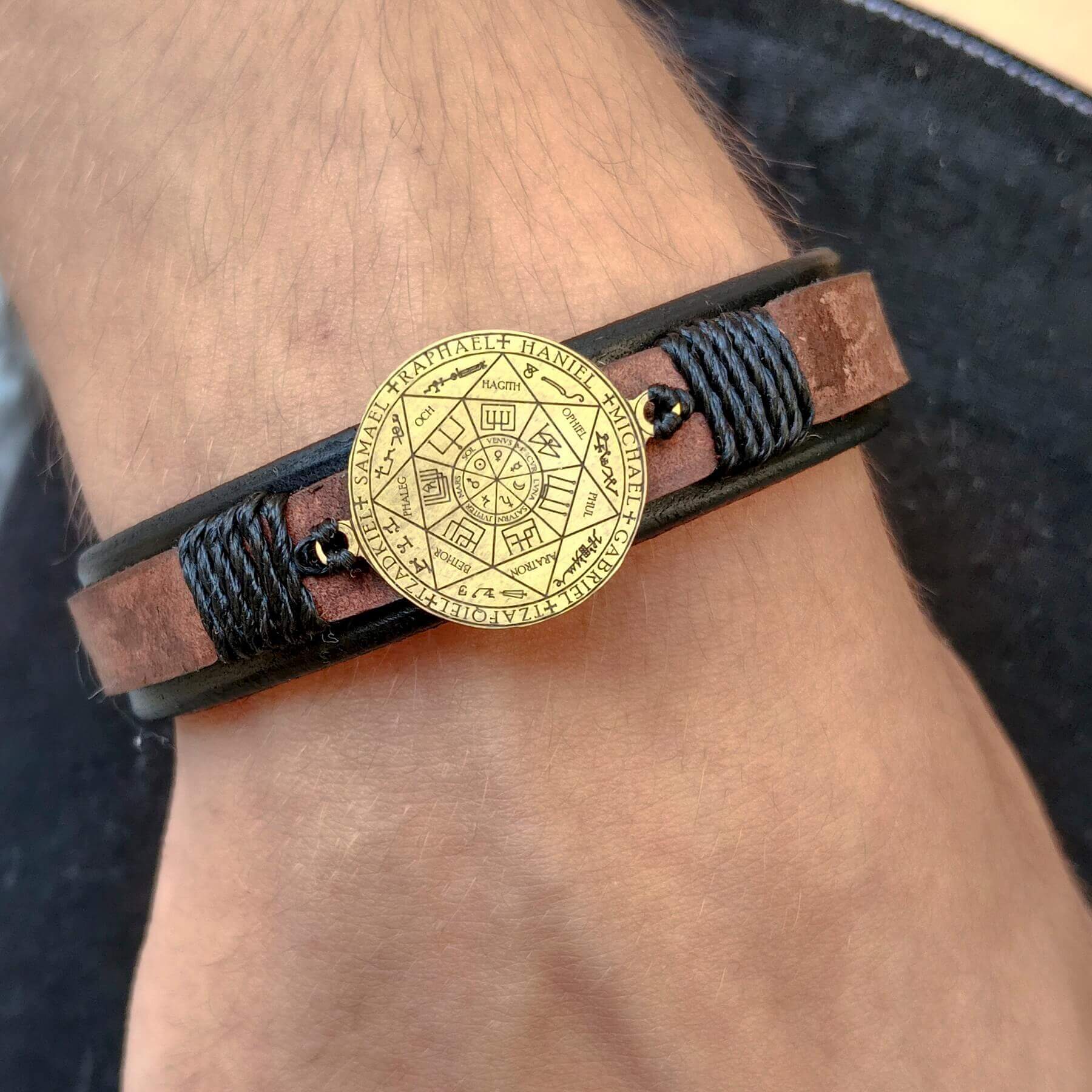 A close-up of a person's wrist wearing a black and brown leather wrap bracelet with black cord accents and a circular gold-toned 7 Archangels Seal pendant.
