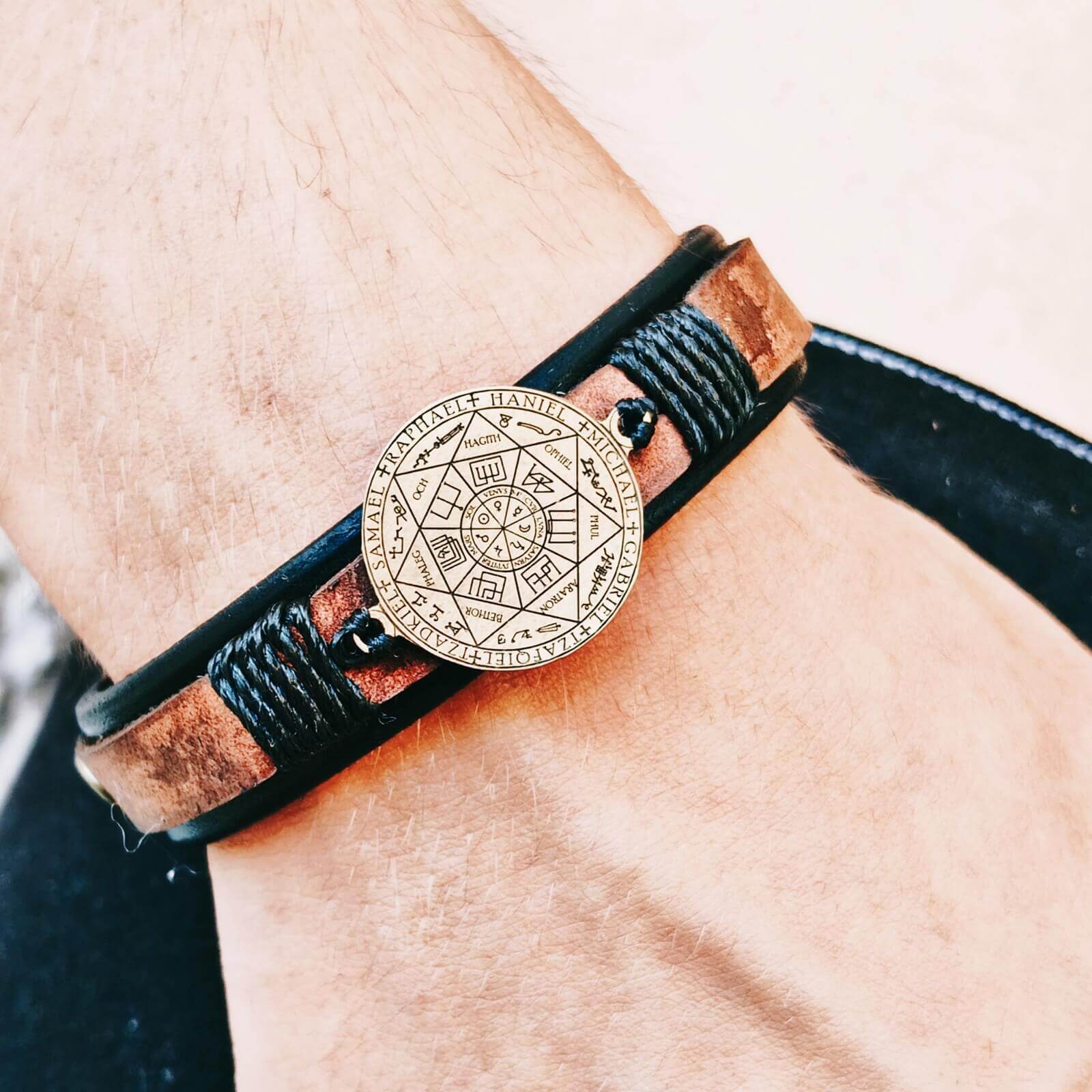 A close-up of a wrist wearing a black wrap leather bracelet with a circular gold-tone 7 Archangels Seal brass pendant.