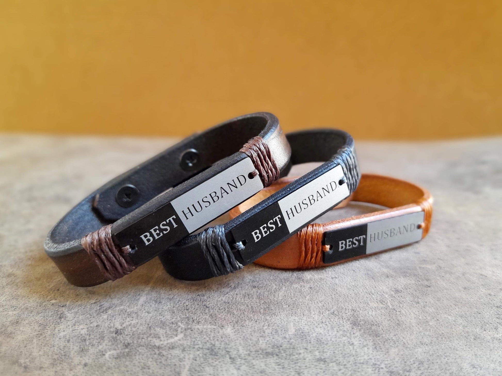 Three stacked leather bracelets with laser engraved stainless steel plates, featuring the words 'BEST' in black and 'HUSBAND' in silverish white, on a textured background.