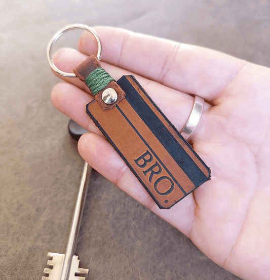 Hand holding a leather keychain with 'BRO.' engraving on a wooden surface