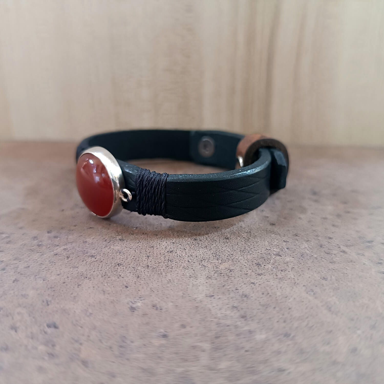 Black leather bracelet with a red stone and silver clasp on a textured surface.
