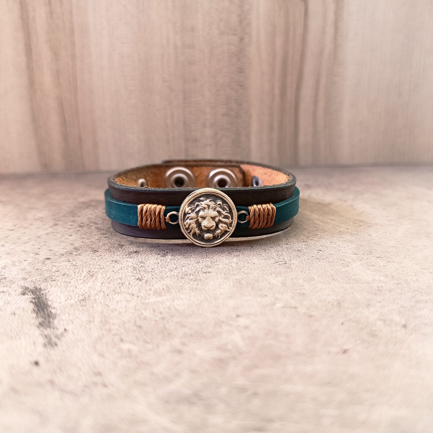 Lion's Quiet Roar Bracelet from Quiet Rebirth Collection. Handcrafted oxidized brass male lion head charm on layered dark brown and petrol blue Crazy Horse leather, laid flat.