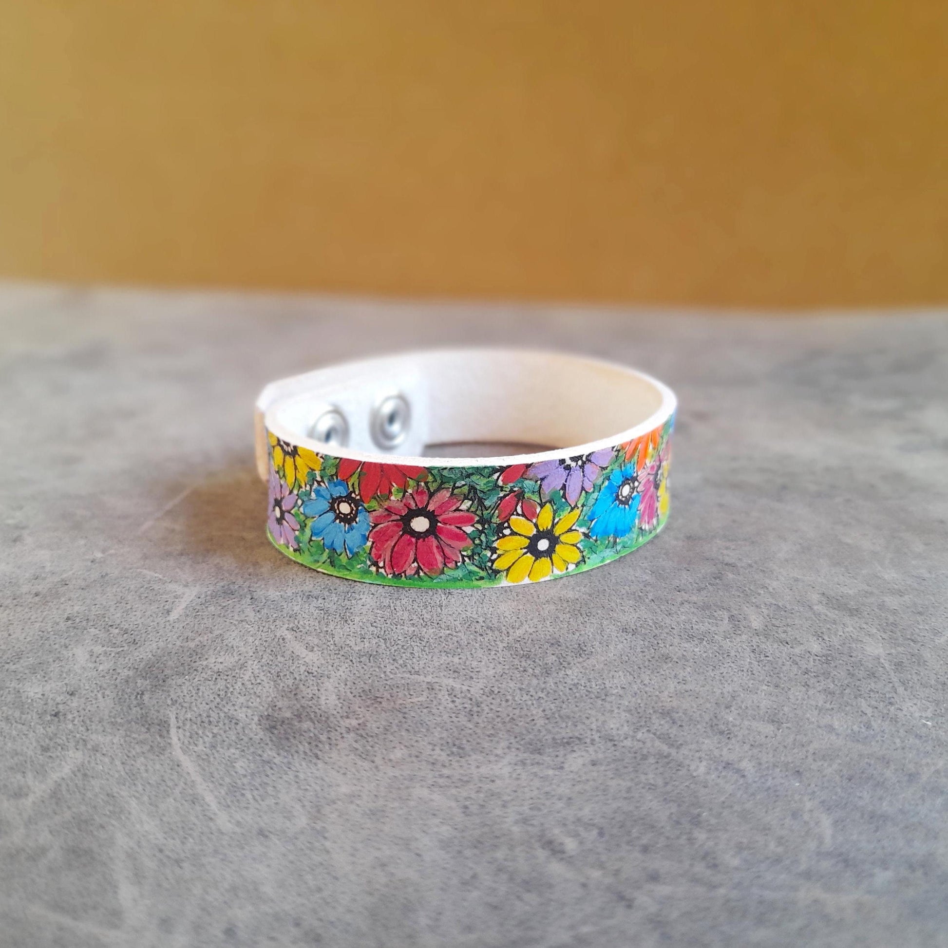 A hand-painted bracelet with colorful spring flowers design, made of creamy white vachetta leather, displayed on a textured background.