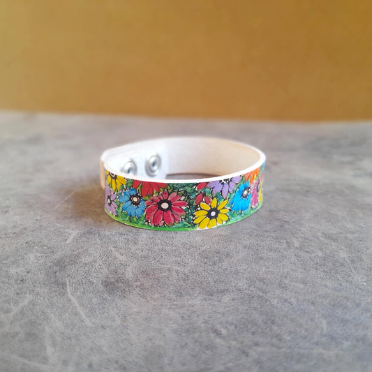 A hand-painted bracelet with colorful spring flowers design, made of creamy white vachetta leather, displayed on a textured background.