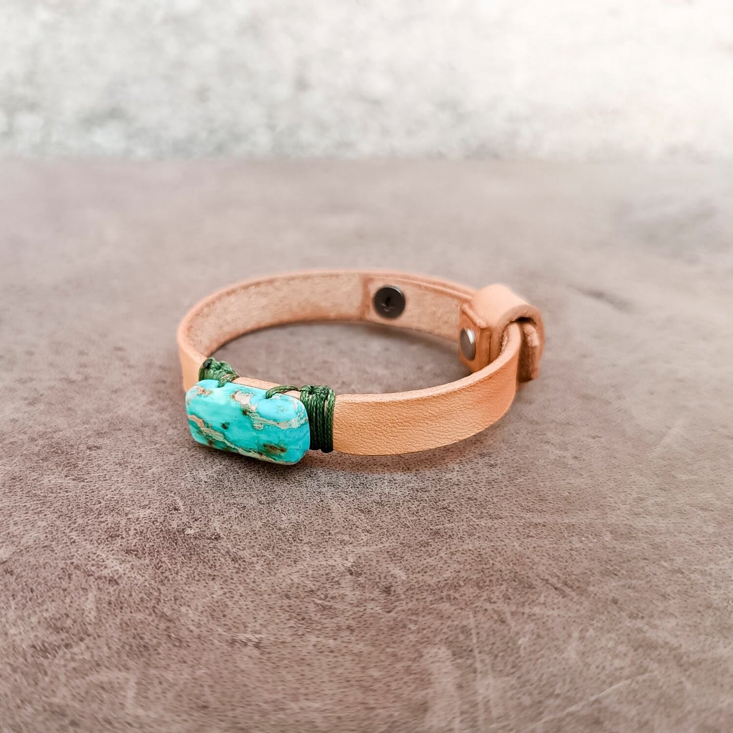 A light tan leather bracelet featuring a central Imperial Jasper stone wrapped with dark green cord, resting on a textured brown surface.