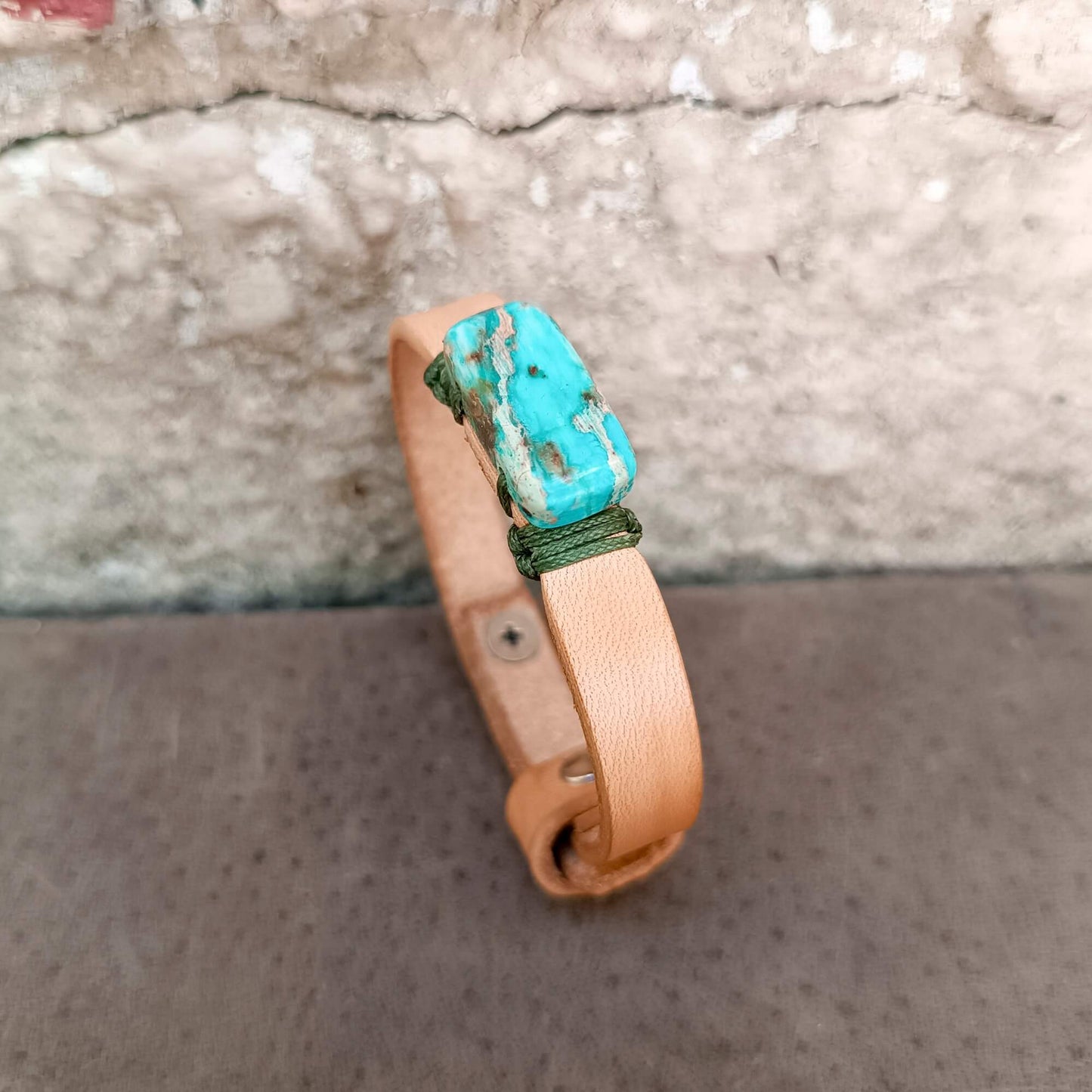 A handmade leather cuff bracelet with a prominent Imperial Jasper stone and green cord wrap, displayed on a textured gray surface against a stone wall.