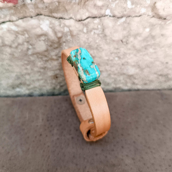 A handmade leather cuff bracelet with a prominent Imperial Jasper stone and green cord wrap, displayed on a textured gray surface against a stone wall.