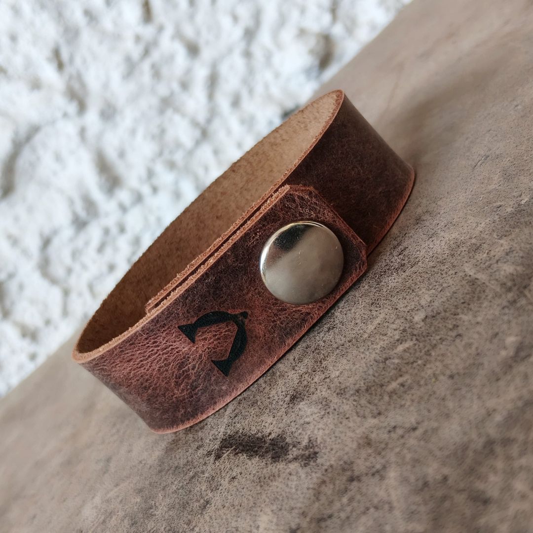 A distressed brown leather cuff bracelet with a silver metal snap closure and a black engraved symbol of Alpha and Omega, resting on a textured gray surface.