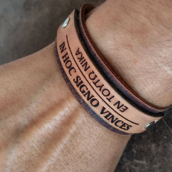 A close-up of a person's wrist wearing a tan and drak brown layered leather bracelet engraved with the phrase 'In Hoc Signo Vinces' in latin and greek.