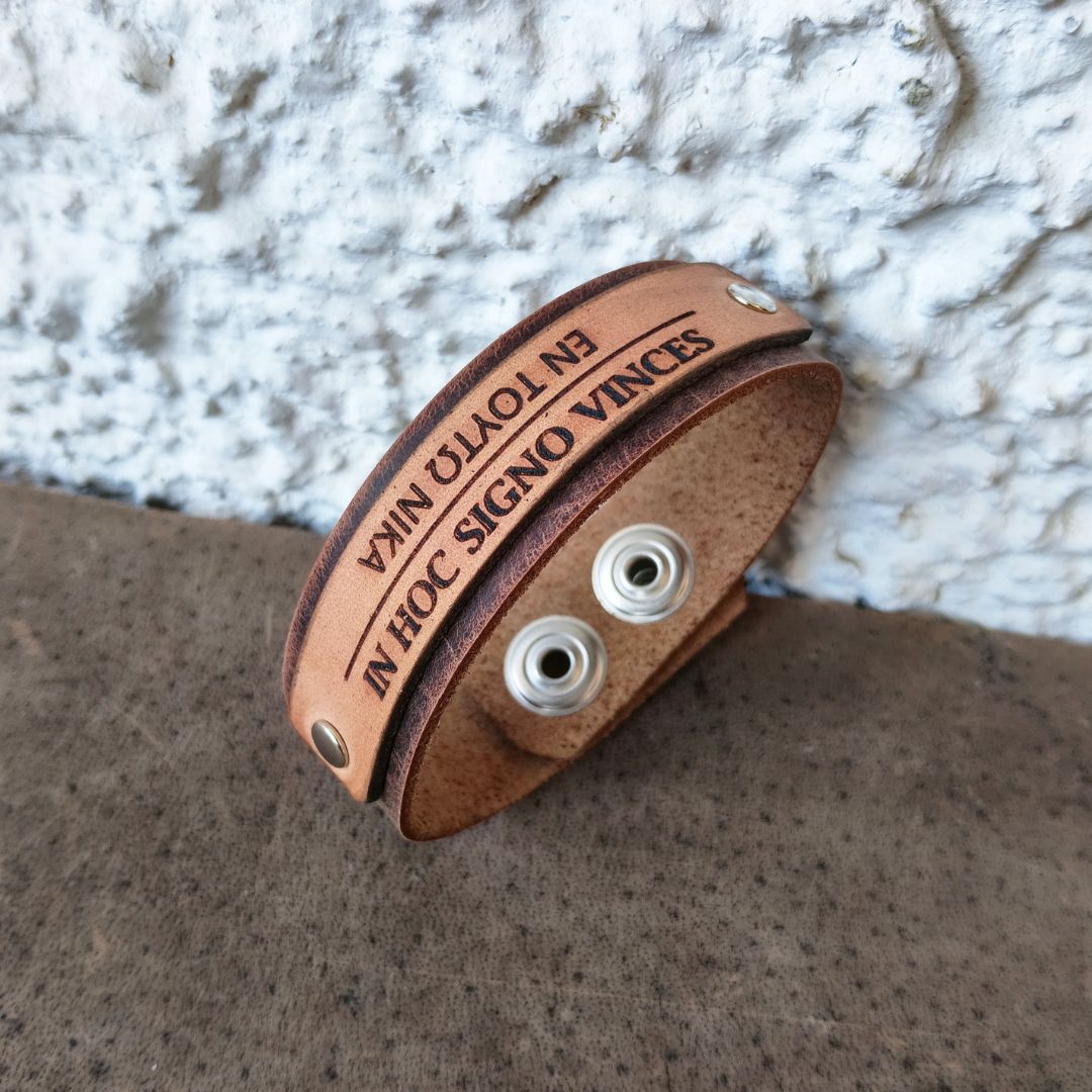 In Hoc Signo Vinces bracelet angle view, photographed resting on a textured brown surface against a white stucco wall. 