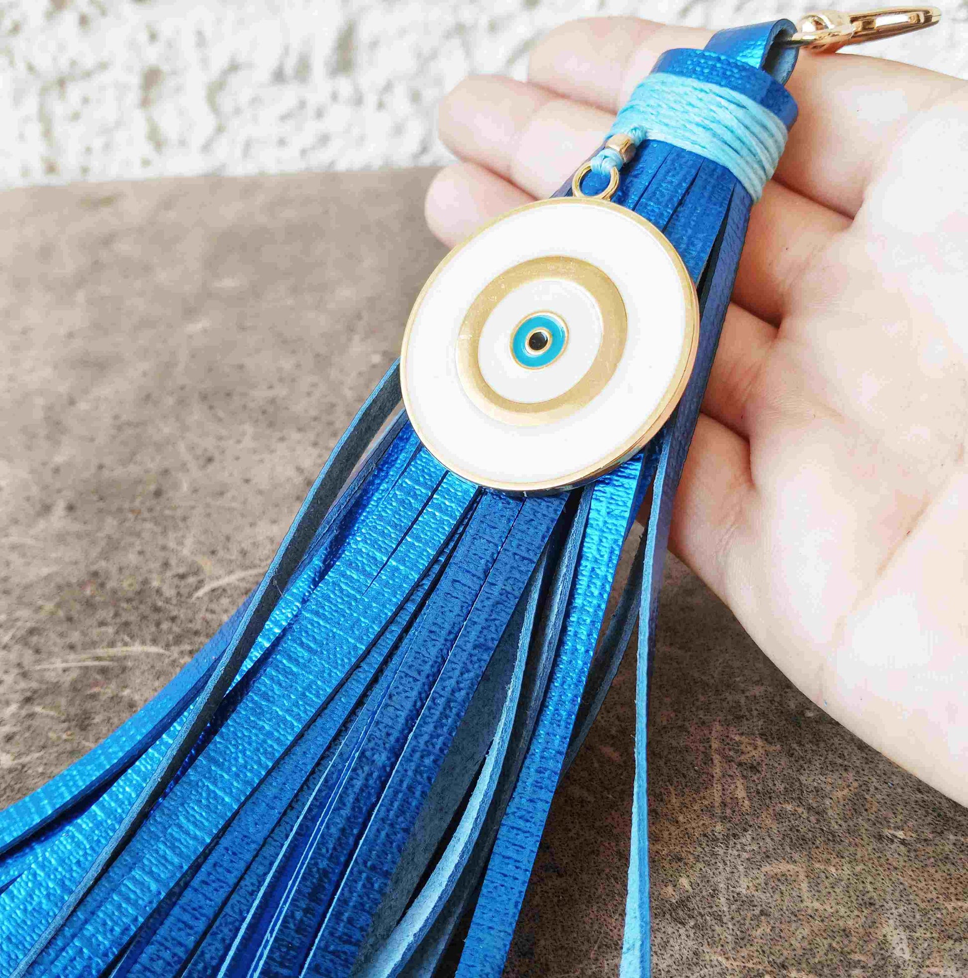 A blue leather tassel keychain or bag charm with a circular evil eye pendant, held in a hand against a textured brown surface.