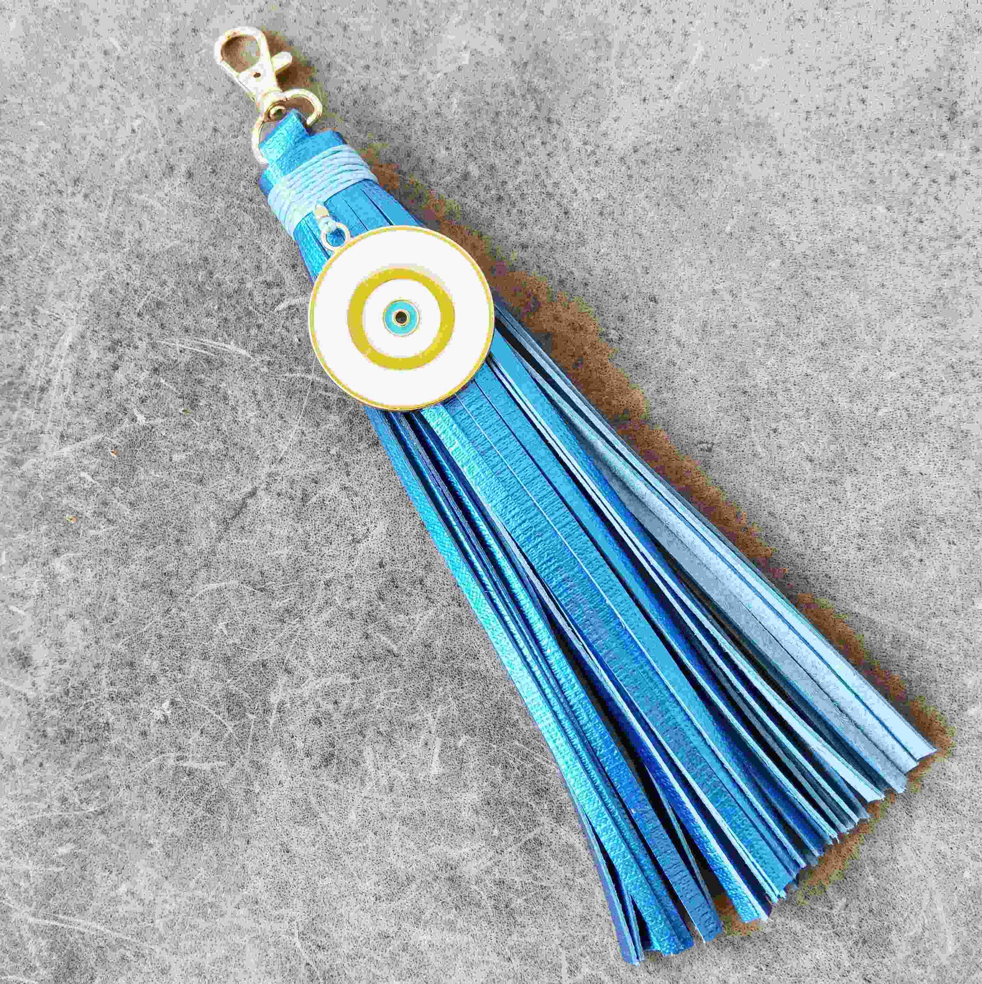 A blue leather tassel keychain with a white evil eye charm featuring gold and blue concentric circles, attached to a gold-tone lobster clasp, set against a textured gray background.