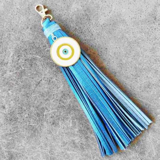 A blue leather tassel keychain with a white evil eye charm featuring gold and blue concentric circles, attached to a gold-tone lobster clasp, set against a textured gray background.