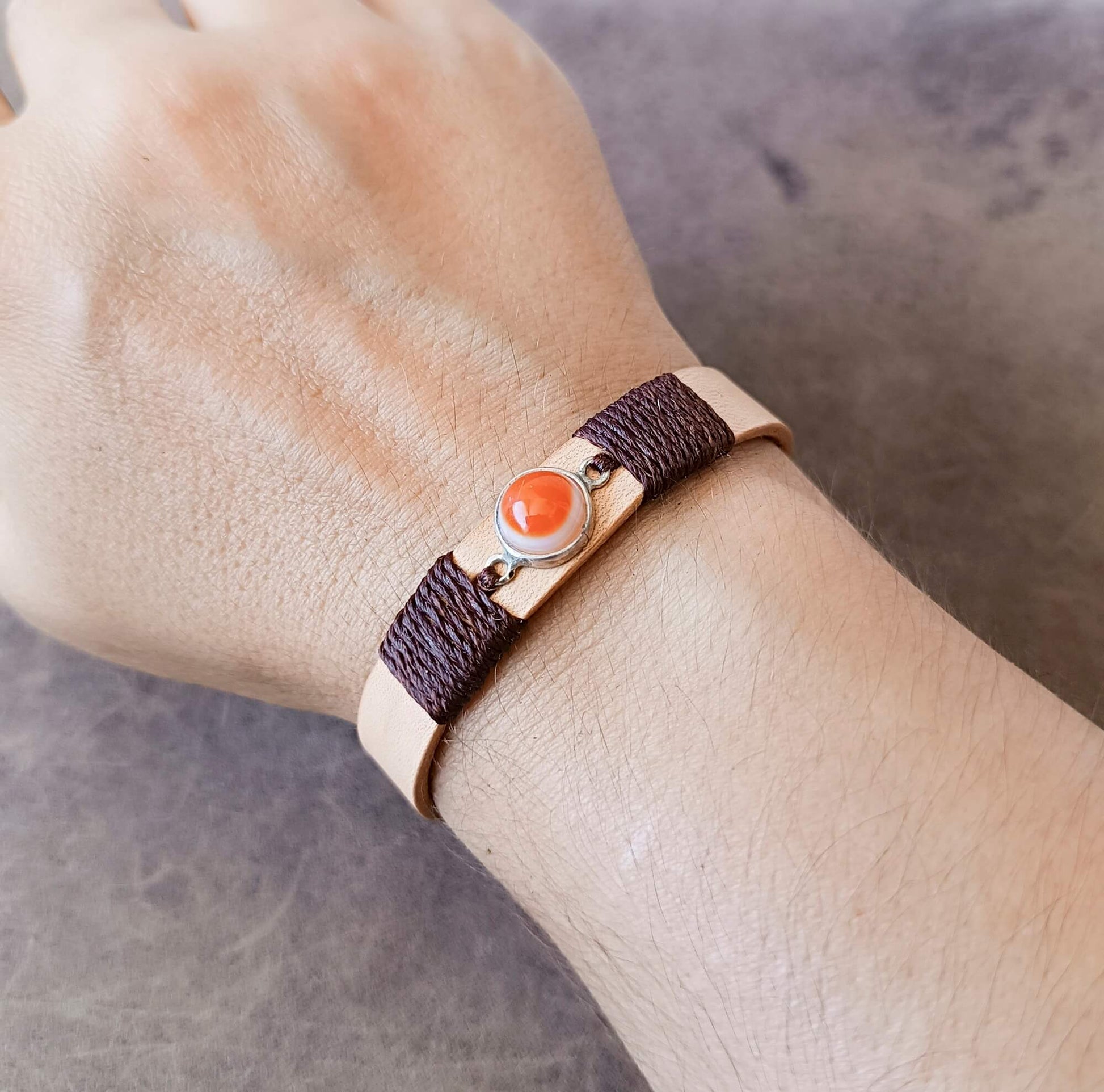 A close-up of a person's wrist wearing a bracelet with a dark brown braided band, a light beige metal band, and a central round orange Carnelian Agate stone in a silver-toned setting.