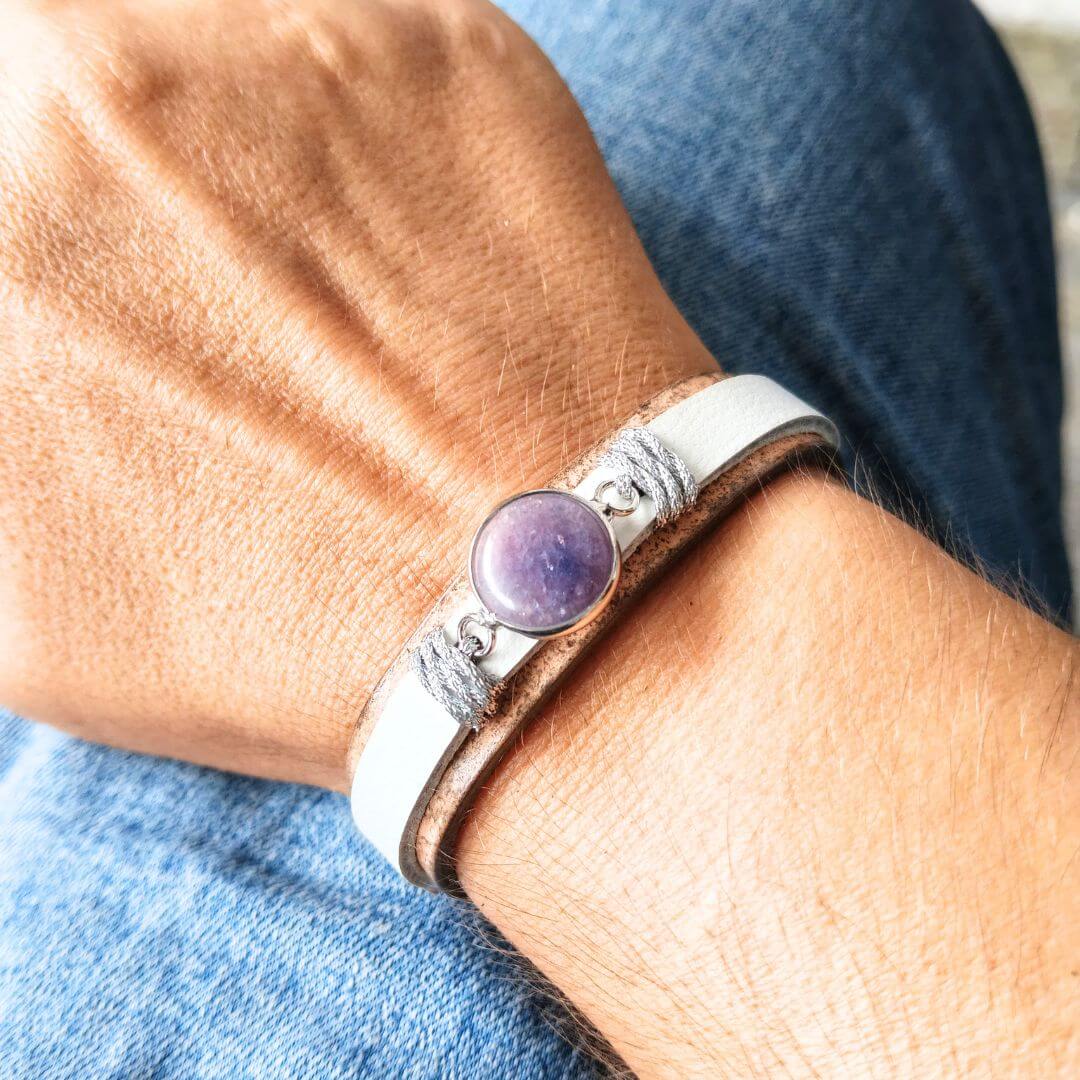 A close-up of a person's wrist wearing a white and tan leather cuff bracelet with a round amethyst gemstone and silver-toned metal details, set against blue denim jeans.