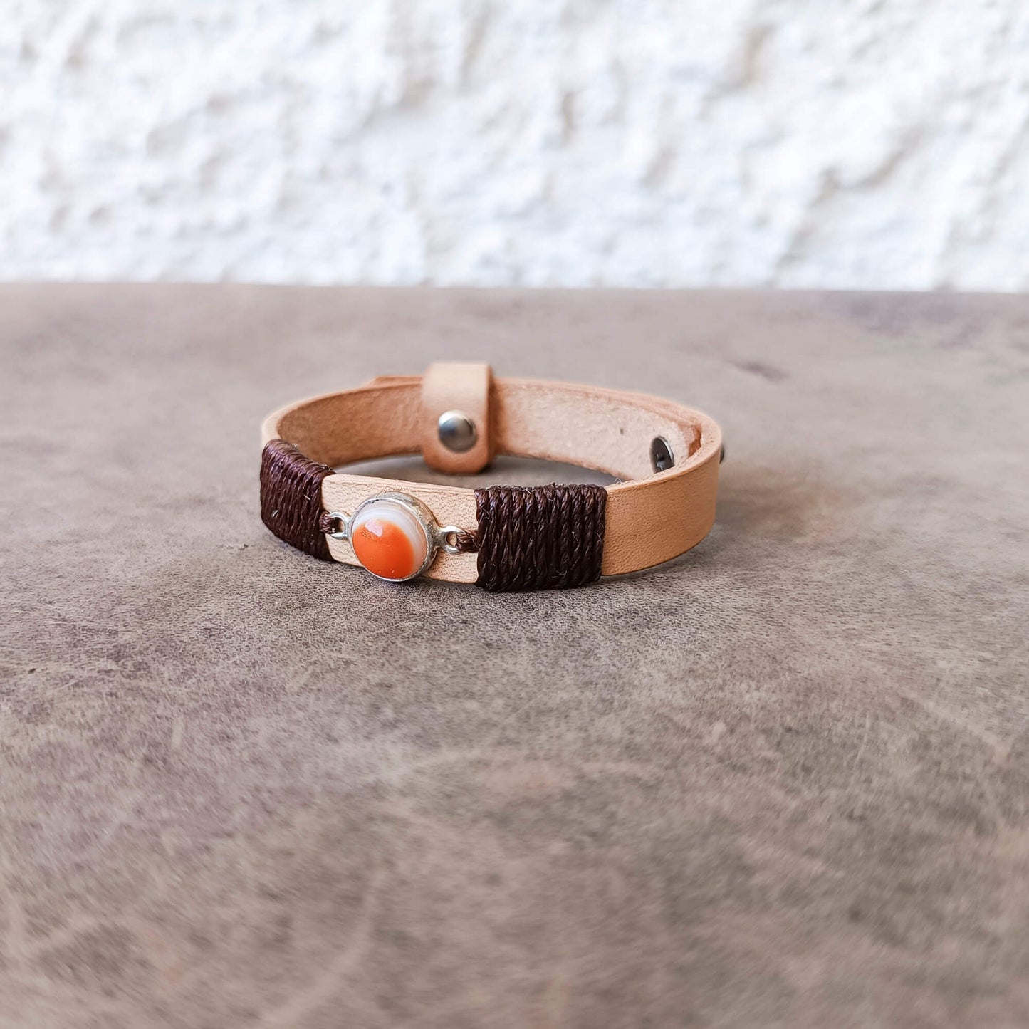 A handmade leather wrap bracelet with dark brown braided cord, a light tan leather band, and a central orange Carnelian Agate stone in a silver bezel, resting on a textured gray surface.