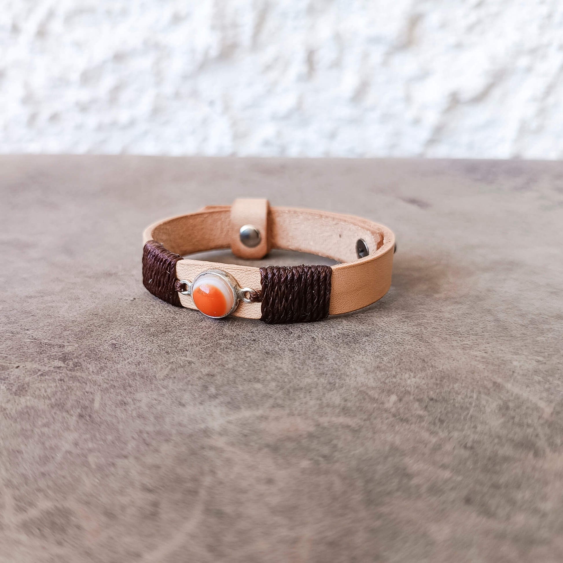A handmade leather wrap bracelet with dark brown braided cord, a light tan leather band, and a central orange Carnelian Agate stone in a silver bezel, resting on a textured gray surface.