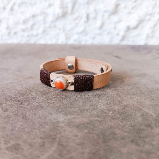 A handmade leather wrap bracelet with dark brown braided cord, a light tan leather band, and a central orange Carnelian Agate stone in a silver bezel, resting on a textured gray surface.