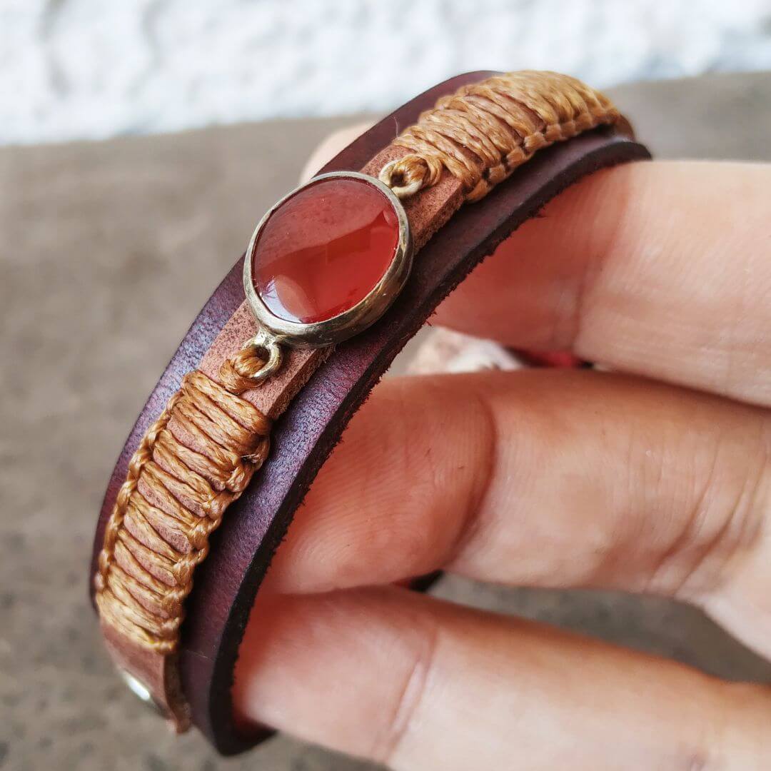A close-up of a hand holding a double-strand leather bracelet with a central oval red stone and braided tan cord details.