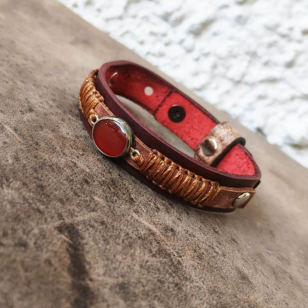 A handmade leather bracelet with a red-orange carnelian cabochon stone, wrapped in copper-colored cord, and resting on a textured brown stone surface.
