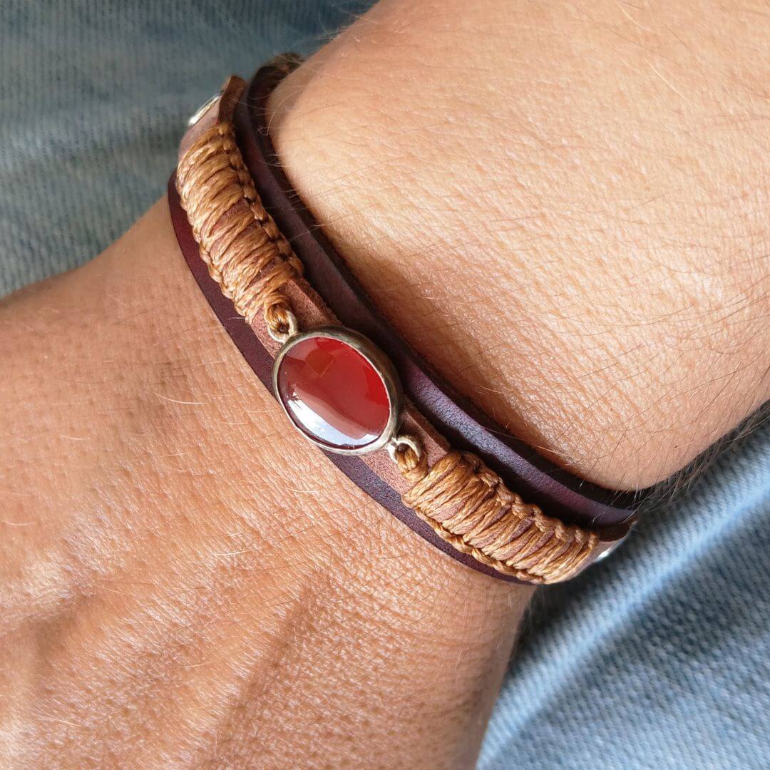 A close-up of a person's wrist wearing a multi-strand bracelet with dark brown leather, tan braided cord, and a central oval red carnelian stone in a gold-toned bezel.