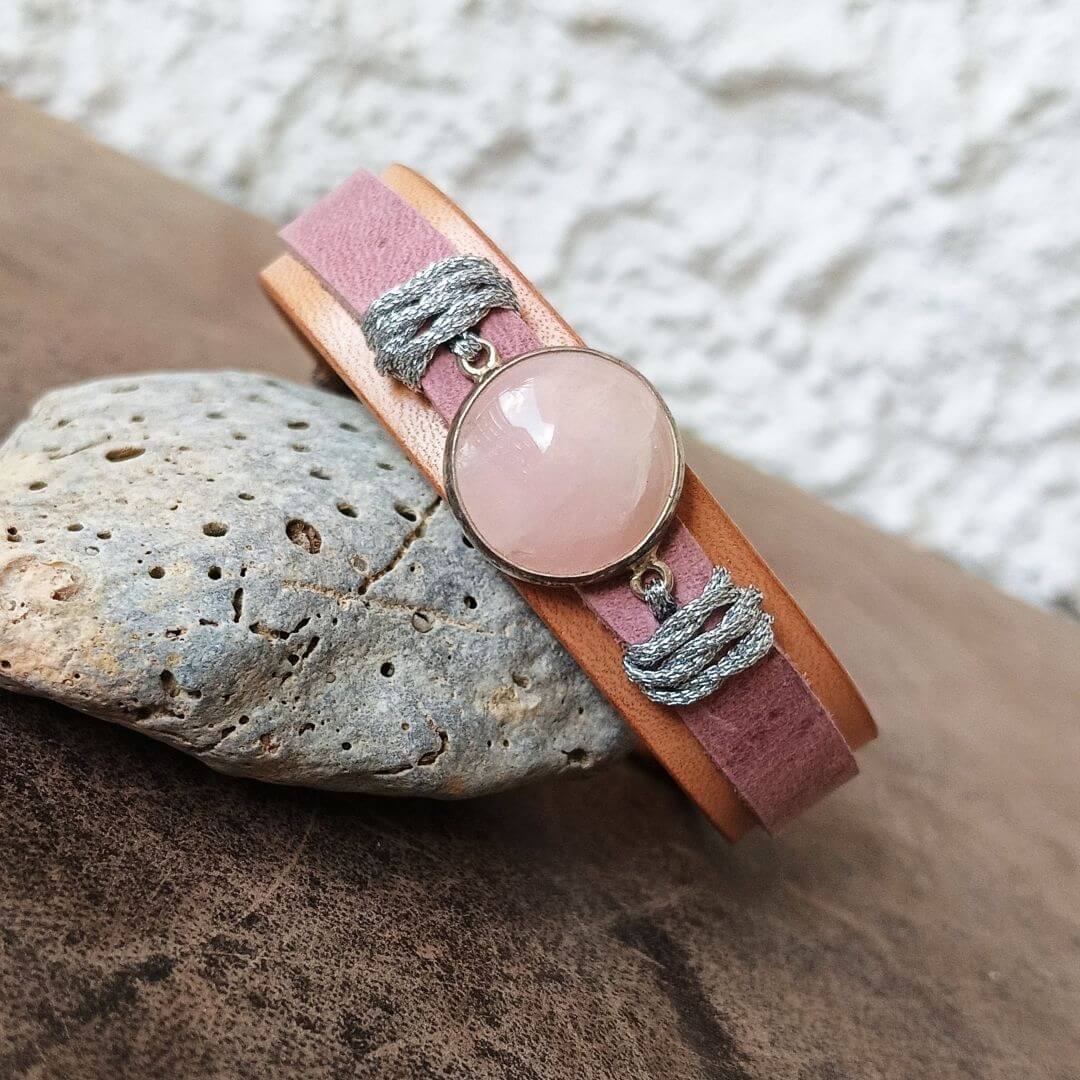 A pink leather wrap bracelet with a round rose quartz stone and gray braided cord accents, displayed on a textured stone over a dark wooden surface.