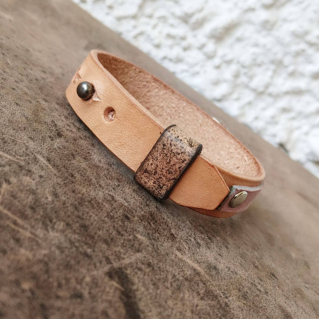 A light tan leather bracelet with a textured dark brown accent tab and brass-tone metal rivets, resting on a rustic brown wooden surface.
